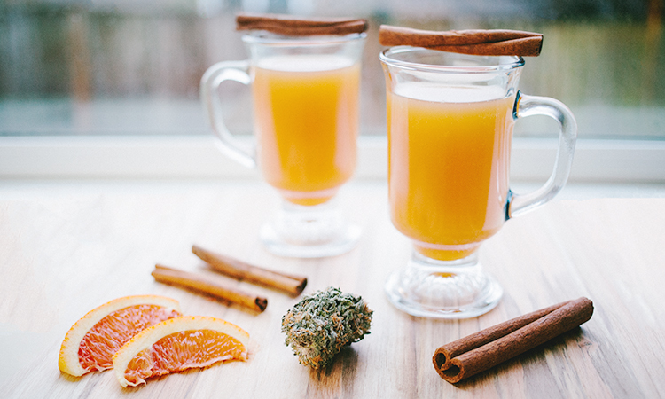 Apple cider infused with cannabis on a wooden table with cinnamon sticks, orange wedges, and cannabis flower
